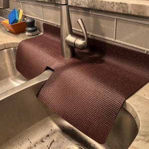 May include: A burgundy sink mat with a textured surface is placed around a stainless steel faucet. The mat is designed to protect the countertop from water damage. A stainless steel sink is in the foreground.