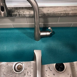 May include: A teal sink mat with a textured surface, placed beneath a stainless steel tap. The mat is designed to catch water and protect the work surface. The sink is made of stainless steel.