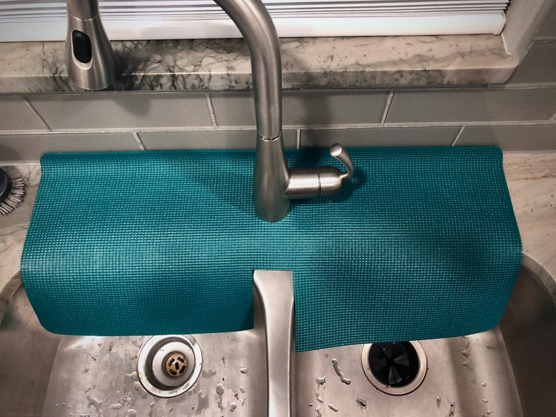 May include: A teal sink mat with a textured surface, placed beneath a stainless steel tap. The mat is designed to catch water and protect the work surface. The sink is made of stainless steel.