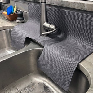May include: A gray sink mat with a textured surface is placed over a stainless steel double sink. The mat is designed to protect the sink from scratches and damage. The mat is placed under a silver faucet.