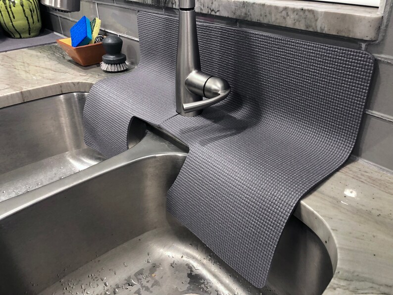 May include: A gray sink mat with a textured surface is placed over a stainless steel double sink. The mat is designed to protect the sink from scratches and damage. The mat is placed under a silver faucet.