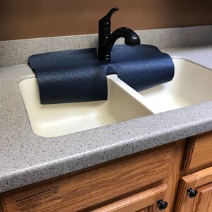Puede incluir: Un fregadero de cocina doble blanco con un grifo negro y una esterilla de yoga azul colocada sobre el fregadero. El fregadero est&aacute; instalado en una encimera con una superficie gris moteada. La encimera est&aacute; unida a armarios de madera con pomos negros.