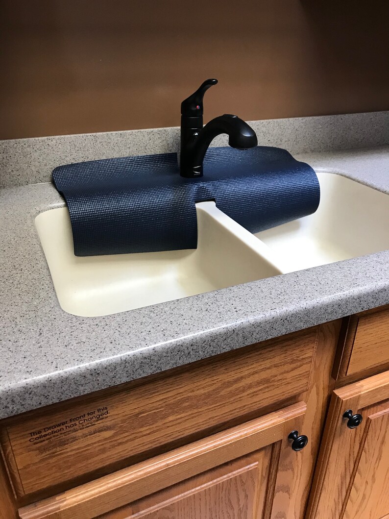 Puede incluir: Un fregadero de cocina doble blanco con un grifo negro y una esterilla de yoga azul colocada sobre el fregadero. El fregadero est&aacute; instalado en una encimera con una superficie gris moteada. La encimera est&aacute; unida a armarios de madera con pomos negros.