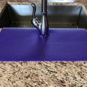May include: A pair of purple sink mats, designed to fit around a faucet, are placed on a speckled granite countertop. The mats are made of a textured material and are positioned in front of a stainless steel sink.