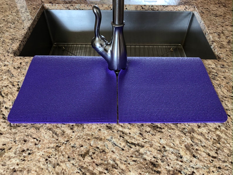 May include: A pair of purple sink mats, designed to fit around a faucet, are placed on a speckled granite countertop. The mats are made of a textured material and are positioned in front of a stainless steel sink.