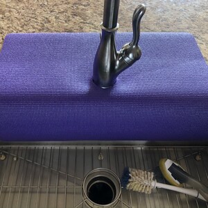 May include: A purple yoga mat placed over a stainless steel kitchen sink. A brushed nickel faucet is centered on the mat. A sink strainer, a scrub brush, and a sponge are visible in the sink.
