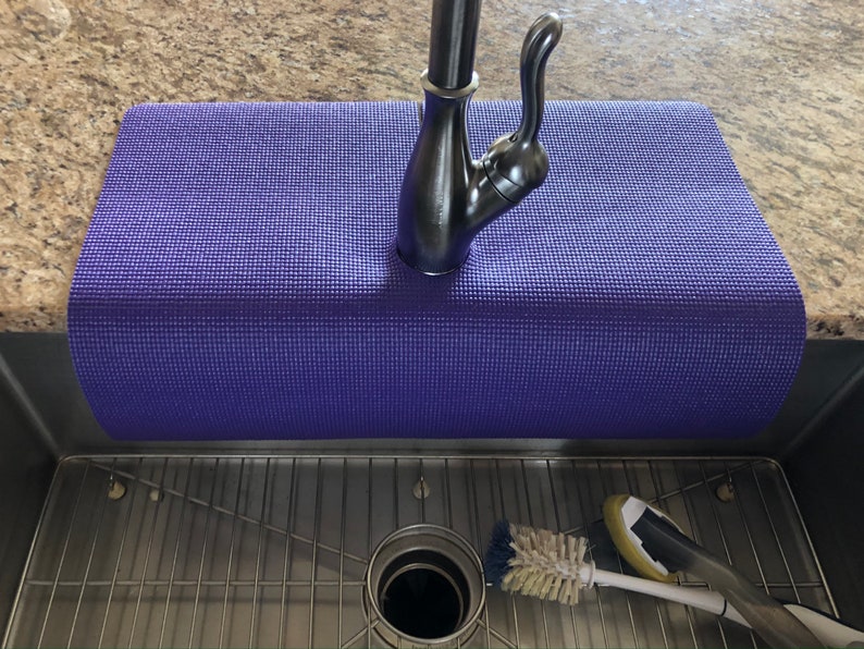 May include: A purple yoga mat placed over a stainless steel kitchen sink. A brushed nickel faucet is centered on the mat. A sink strainer, a scrub brush, and a sponge are visible in the sink.