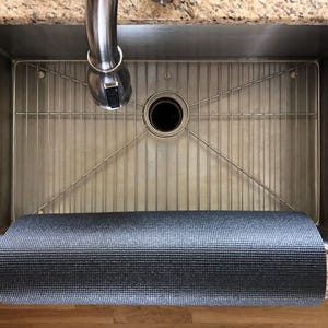 May include: A stainless steel kitchen sink with a metal grid and a rolled-up gray yoga mat. The sink has a faucet and is set into a granite countertop. The yoga mat is rolled up at the bottom of the sink.