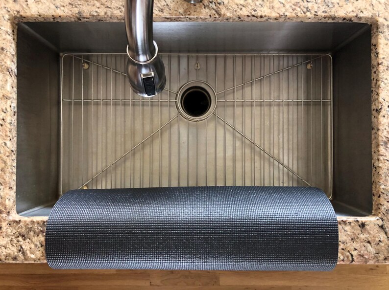 May include: A stainless steel kitchen sink with a metal grid and a rolled-up gray yoga mat. The sink has a faucet and is set into a granite countertop. The yoga mat is rolled up at the bottom of the sink.