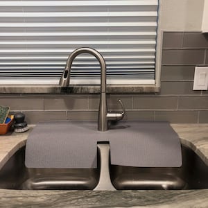May include: Stainless steel kitchen sink with a high-arc faucet. The sink has two basins and a gray mat. The faucet is positioned in front of a window with white blinds. The countertop is a light gray with darker gray veining.