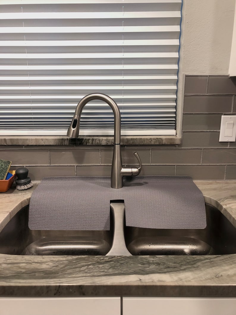 May include: Stainless steel kitchen sink with a high-arc faucet. The sink has two basins and a gray mat. The faucet is positioned in front of a window with white blinds. The countertop is a light gray with darker gray veining.