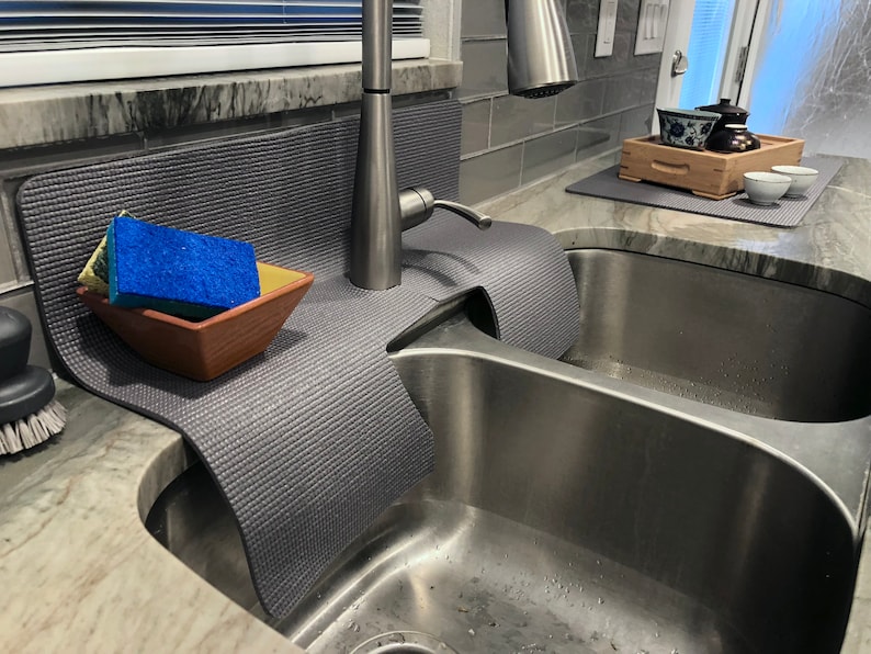 May include: A gray kitchen sink mat with a blue and yellow sponge in a brown ceramic dish. The mat is placed over a stainless steel double sink with a chrome faucet.