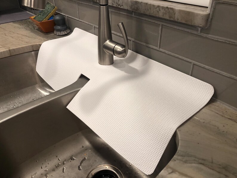 May include: A white, textured sink mat sits over a stainless steel sink. The mat has a cutout for the tap and is designed to protect the sink's surface. The tap is made of brushed nickel.