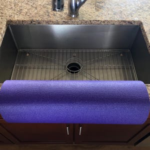 May include: A rolled-up purple yoga mat rests on the edge of a stainless steel kitchen sink. The sink is set into a granite countertop. The mat is a solid color and appears to be made of a textured material. The cabinet doors below the sink are a dark brown color.