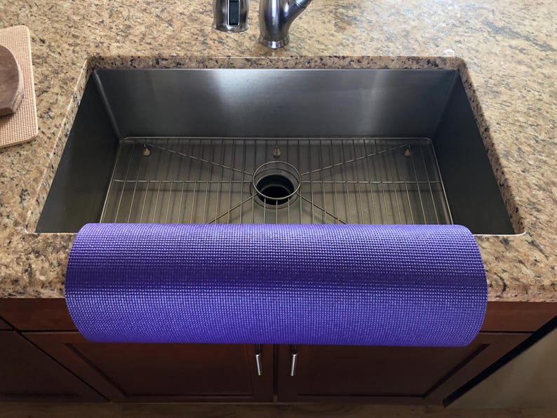 May include: A rolled-up purple yoga mat rests on the edge of a stainless steel kitchen sink. The sink is set into a granite countertop. The mat is a solid color and appears to be made of a textured material. The cabinet doors below the sink are a dark brown color.
