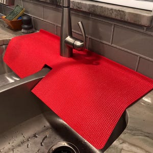 May include: A vibrant red sink mat with a textured surface. The mat is designed to fit around a tap and protect the work surface from water. The mat is placed over a stainless steel sink.