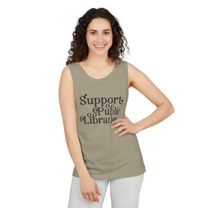 May include: A sage green sleeveless tank top featuring the dark brown text "Support Public Libraries" printed in a stylized, rounded font across the chest. The garment has a scoop neckline and a straight hemline, providing a simple, casual look.