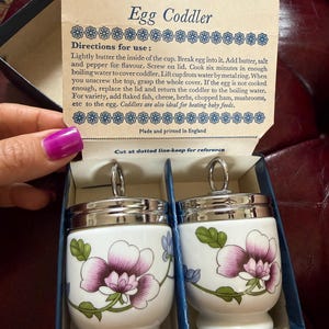 May include: Two Royal Worcester egg coddlers in a box with a lid featuring instructions. The white porcelain coddlers have a floral pattern with purple and pink petals, green leaves, and blue accents. Each has a silver-colored metal lid with a ring.
