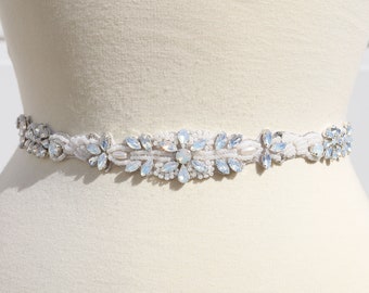 May include: A white beaded bridal sash with a large, silver, and light blue rhinestone embellishment.