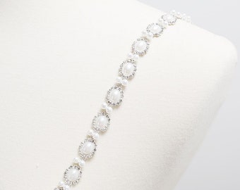 May include: A white pearl and rhinestone trim with a silver chain. The trim is displayed on a white mannequin.