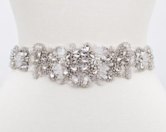 May include: A decorative belt featuring an intricate arrangement of clear crystals, rhinestones, and small white pearls set against a silver-toned base. The embellishments form a symmetrical, ornate pattern that spans the width of the belt.