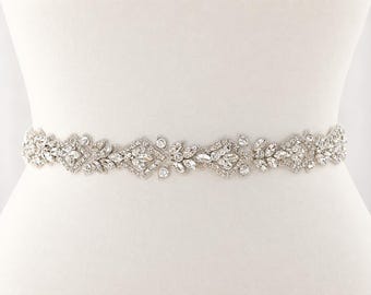 May include: A silver-toned belt featuring an intricate arrangement of clear, faceted crystals and rhinestones. The design consists of repeating geometric and leaf-like patterns, creating a continuous, sparkling band of embellishments across the front.
