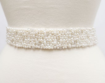 May include: A white beaded sash with a pearl-like design. The sash is made of a white fabric and is decorated with small, white beads that are arranged in a random pattern.