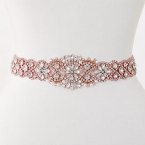 May include: A rose gold belt featuring an intricate arrangement of clear crystals and small white pearls. The design consists of a central floral-inspired cluster flanked by repeating scalloped patterns, all set in a metallic rose gold frame.