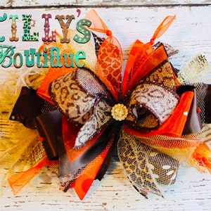 May include: A large, multi-layered bow made with brown, orange, and gold ribbon. The bow has a leopard print pattern and a sparkly finish. The bow is tied with a brown ribbon and has a small, gold button in the center. The bow is on a white wooden background. Tilly's Boutique is written in the top left corner of the image.