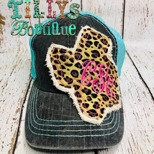 May include: A black and turquoise trucker hat with a leopard print Texas-shaped patch and pink monogram. The hat has a mesh back and a distressed look.