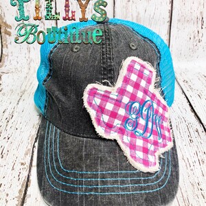 Op de afbeelding: Een zwarte en blauwe trucker-pet met een roze en witte gingham Texas-vormige patch met een monogram. De pet heeft een blauwe mesh-rug en een zwarte klep.