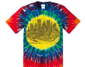 Puede incluir: Una camiseta tie-dye con un círculo amarillo en el centro que presenta una ilustración en blanco y negro del horizonte de Pittsburgh. El texto "PITTSBURGH" está impreso debajo de la ilustración.