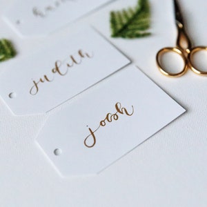 May include: White gift tags with gold calligraphy names, including "Josh" and "Judith". A pair of gold scissors and sprigs of greenery are arranged on a white surface, creating a clean, elegant aesthetic. Ideal for weddings or special events.