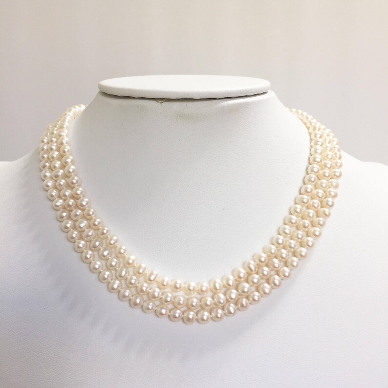 May include: A three-strand pearl necklace displayed on a white mannequin. The pearls are a creamy white color and are evenly sized. The necklace is draped in a gentle curve, showcasing its elegant design. The background is plain white.