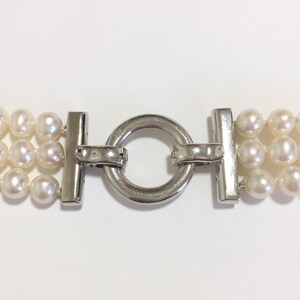 May include: Close-up of a three-strand pearl necklace with a silver clasp. The pearls are white and round, and the clasp is a shiny silver metal. The necklace is displayed against a white background.