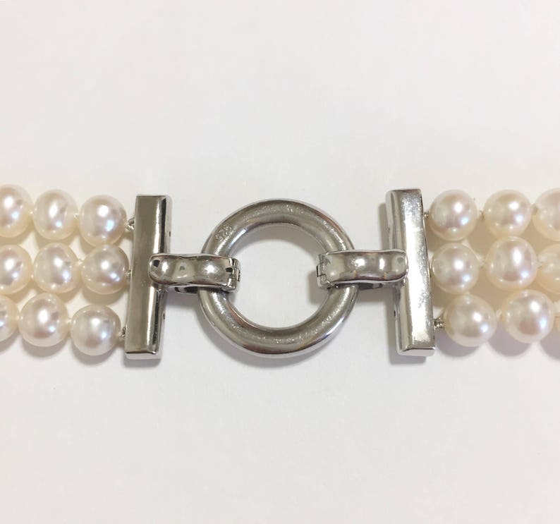 May include: Close-up of a three-strand pearl necklace with a silver clasp. The pearls are white and round, and the clasp is a shiny silver metal. The necklace is displayed against a white background.