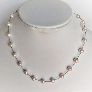 May include: A delicate pearl necklace featuring a strand of luminous, off-white pearls. The pearls are strung on a dark, thin cord, creating a minimalist and elegant design. The necklace is displayed on a white mannequin bust, showcasing its shape and style.