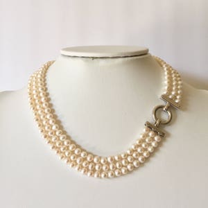 May include: A three-strand pearl necklace with a silver-tone clasp. The pearls are a creamy white color and are evenly sized. The clasp is a simple design with a circular ring.