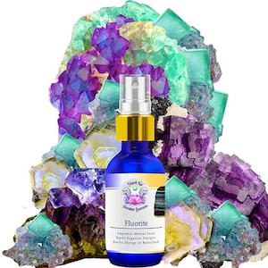 Puede incluir: Una botella de vidrio azul con una boquilla de pulverización dorada y una etiqueta que dice "Fluorite -  Empowers Mental Focus, Repels Negative Energies,  Invites Energy of Balance/Truth". La botella está rodeada de un montón de cristales de fluorita morados, verdes y amarillos.