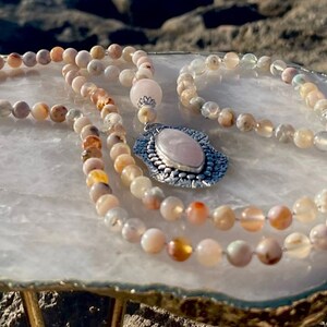 May include: A pink and white opal gemstone mala necklace with a silver pendant featuring a pink rose quartz stone. The necklace is displayed on a white quartz slab.