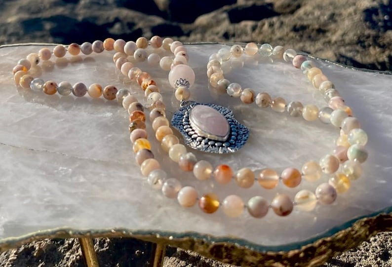 May include: A pink and white opal gemstone mala necklace with a silver pendant featuring a pink rose quartz stone. The necklace is displayed on a white quartz slab.