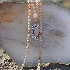May include: A long, multi-strand necklace made of light pink, brown, and white beads. The necklace features a large, oval-shaped pendant with a pink stone set in a silver frame.