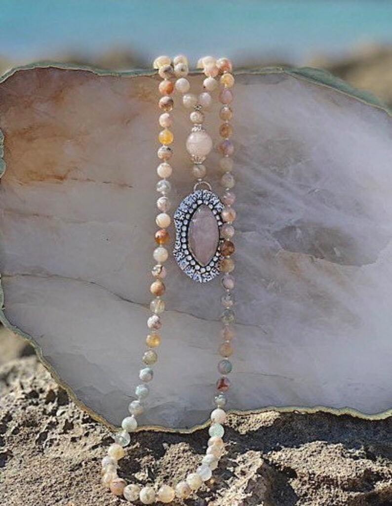 May include: A long, multi-strand necklace made of light pink, brown, and white beads. The necklace features a large, oval-shaped pendant with a pink stone set in a silver frame.