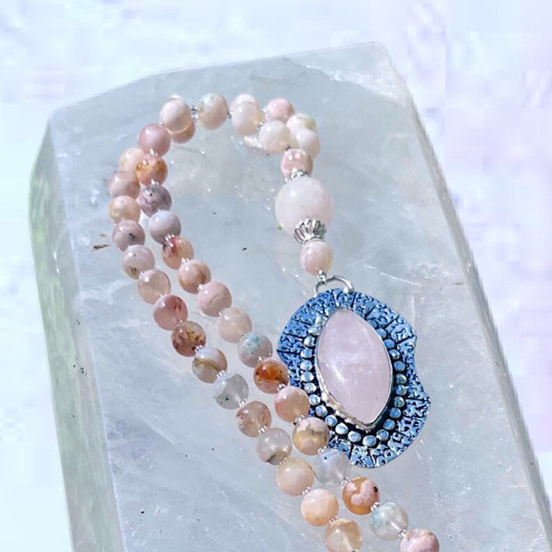 May include: A pink and white gemstone mala necklace with a silver pendant featuring a large rose quartz stone.