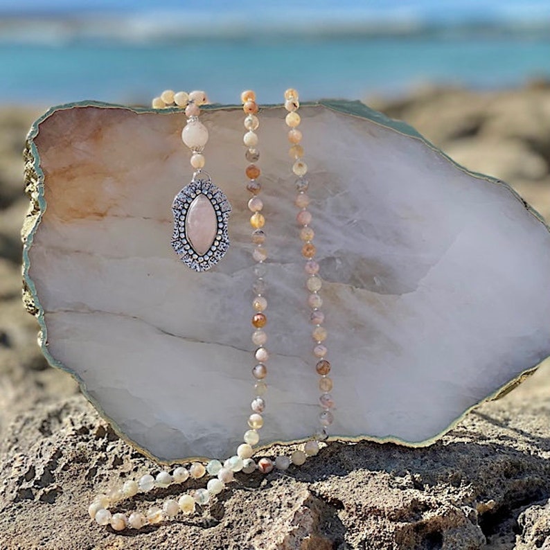 A beaded mala necklace with a light pink rose quartz oval pendant set in ornately designed sterling silver, with light pink, multi-hued beads is draped over a crystal slab on a rock with the ocean behind it.