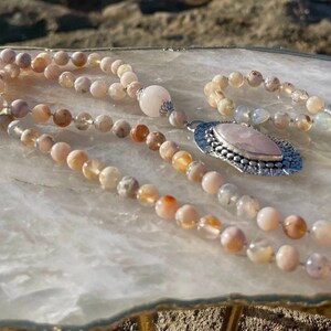 May include: A pink and white agate mala necklace with a silver pendant featuring a rose quartz stone. The necklace is laid out on a white surface.