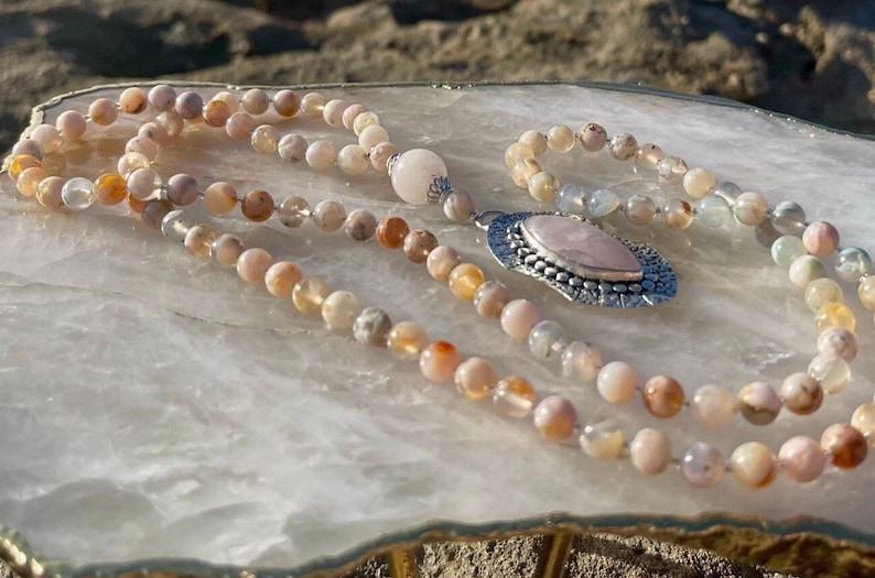 May include: A pink and white agate mala necklace with a silver pendant featuring a rose quartz stone. The necklace is laid out on a white surface.