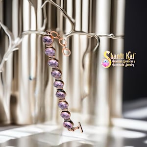 Op de afbeelding: Een armband met een reeks ovaalvormige, lichtpaarse doorschijnende stenen in een koperkleurig metalen montuur. Het sieraad heeft een kleine schakelketting aan de bovenkant en een T-sluiting aan de onderkant. Tekst leest Shanti Kai.