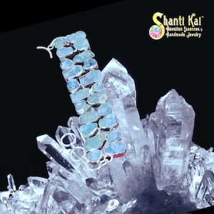 May include: A bracelet featuring multiple rough-cut, pale blue aquamarine stones set in silver metal links. The piece includes a silver toggle clasp and is displayed against a cluster of clear, pointed quartz crystals with the Shanti Kai logo.