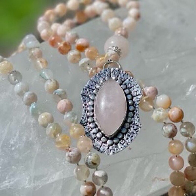 May include: A multi-strand mala necklace with a silver pendant featuring a rose quartz stone. The necklace is made with various shades of pink, green, and brown stones.